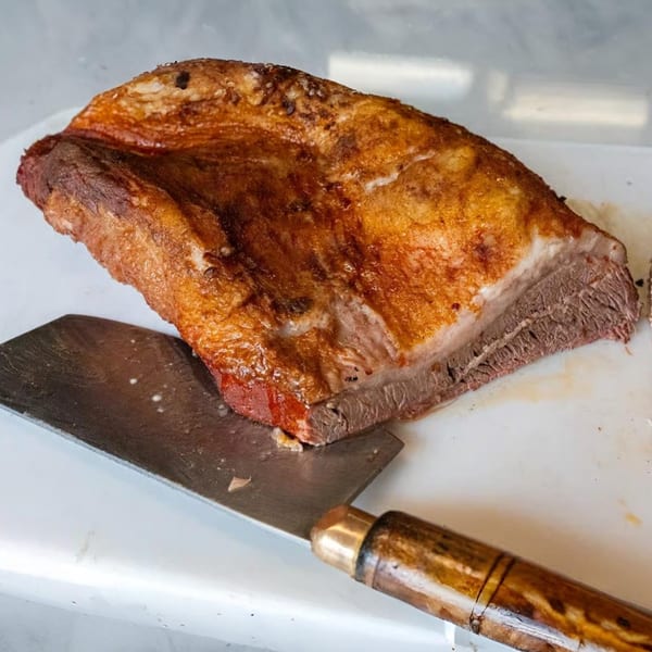 A large, golden-brown cut of Roy's Hickory Pit BBQ brisket rests on a white cutting board next to a meat cleaver.